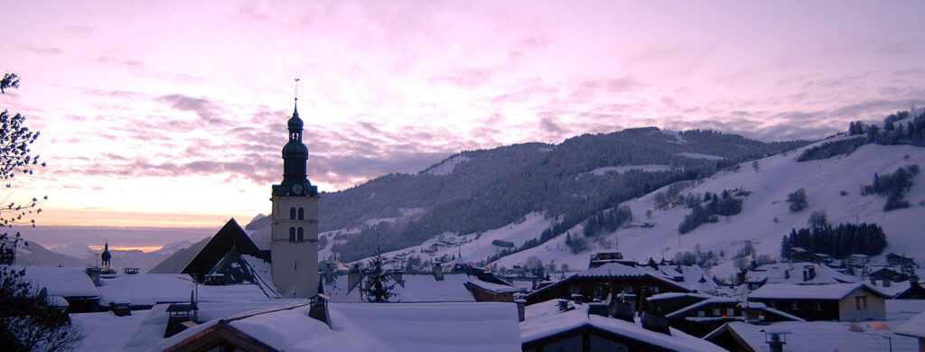 Village de Megève au coucher du soleil avec clocher d'église et montagnes enneigées dans les Alpes françaises.